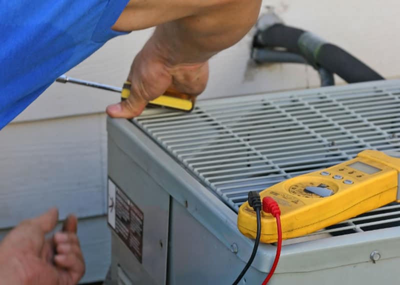 Technician using a screwdriver on an HVAC unit with a multimeter nearby