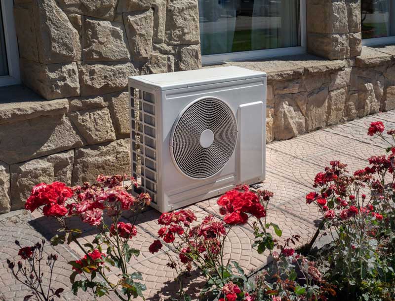 Outdoor mini-split AC unit installed near a stone wall and flower bed